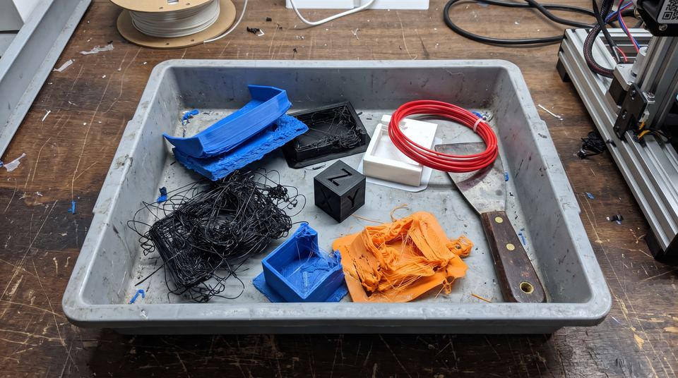Pièces imprimées ratées et réussies rangées dans un bac, avec un cube de test, un bout de filament et une spatule.